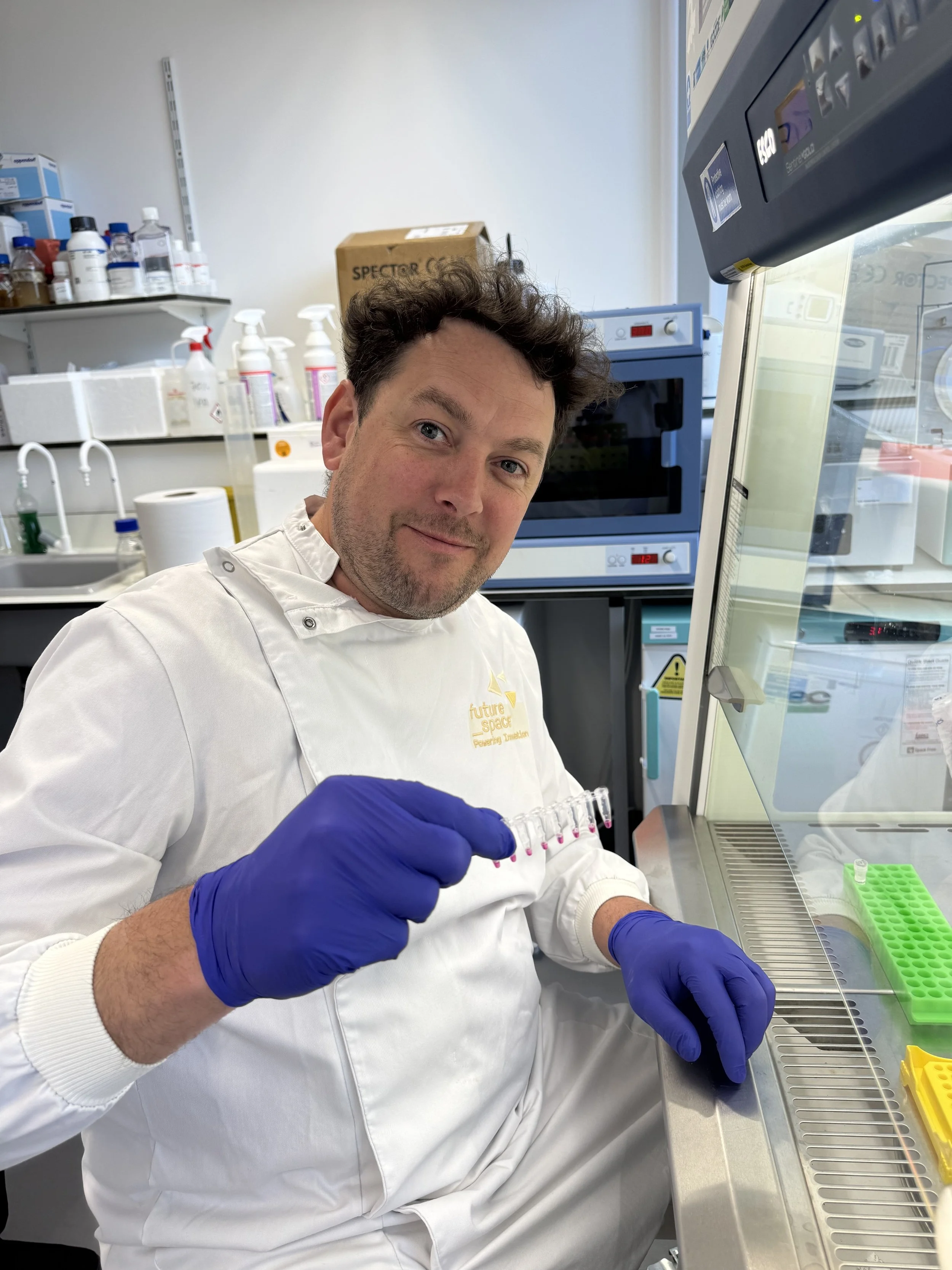 A scientist in a white lab coat and blue gloves working in a laboratory. They are holding a vial with pink liquid and working under a biosafety cabinet. The background shows lab equipment, bottles, and a microwave oven.
