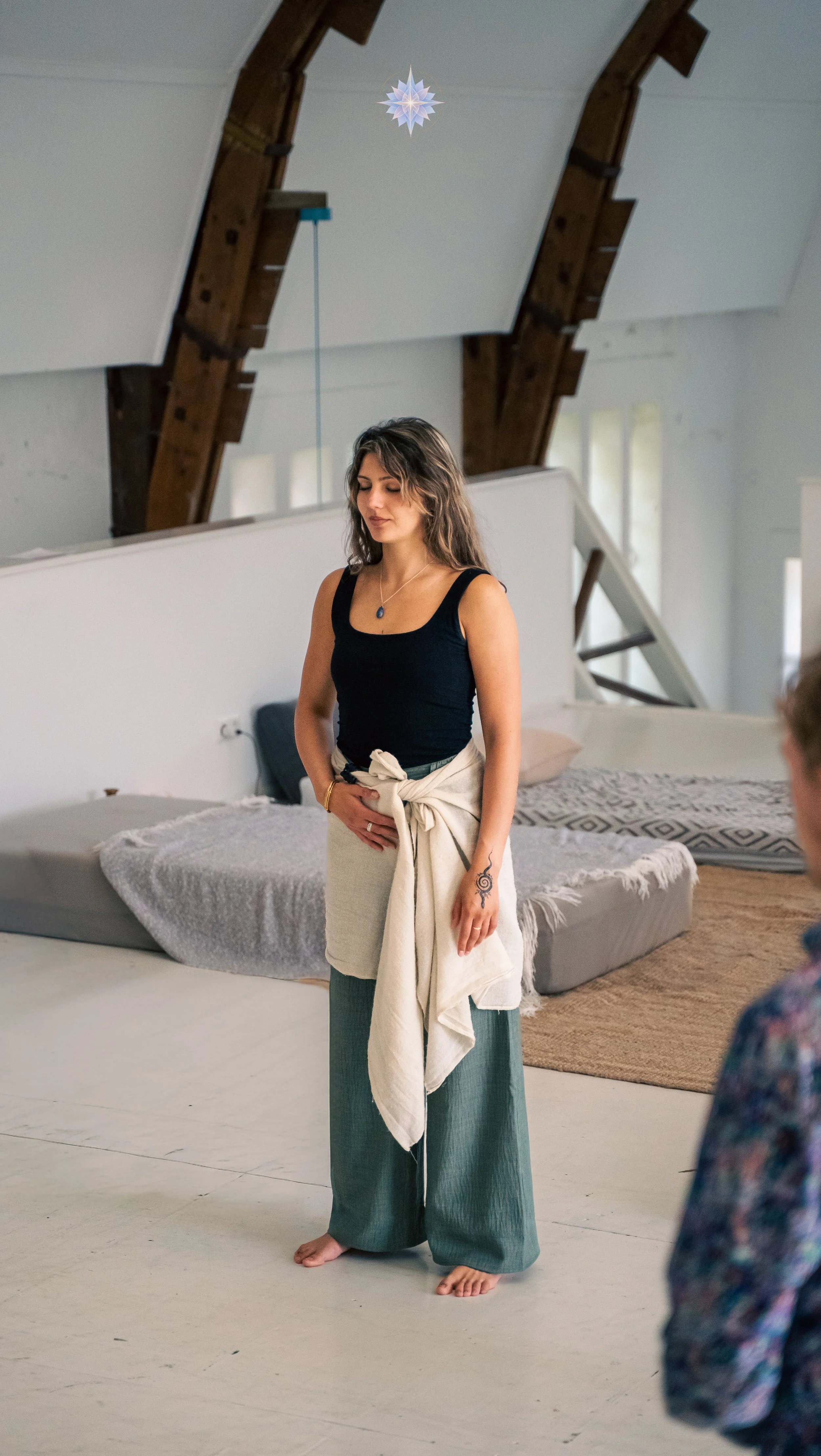 A woman standing barefoot on a light-colored floor in a room with a sloped ceiling, wooden beams, and large windows. She is wearing a black tank top, green wide-leg pants, and has a light-colored jacket tied around her waist. She has wavy brown hair, a necklace, and a tattoo on her left hand. There is a bed with a blanket and pillow behind her, and a person with a floral shirt partially visible in the foreground.