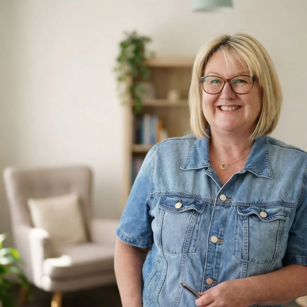 Louise Redding Counselling, Online therapy across the UK. Louise with blonde hair and glasses smiling, wearing a denim jacket, standing indoors in a room with a bookshelf and a beige chair.