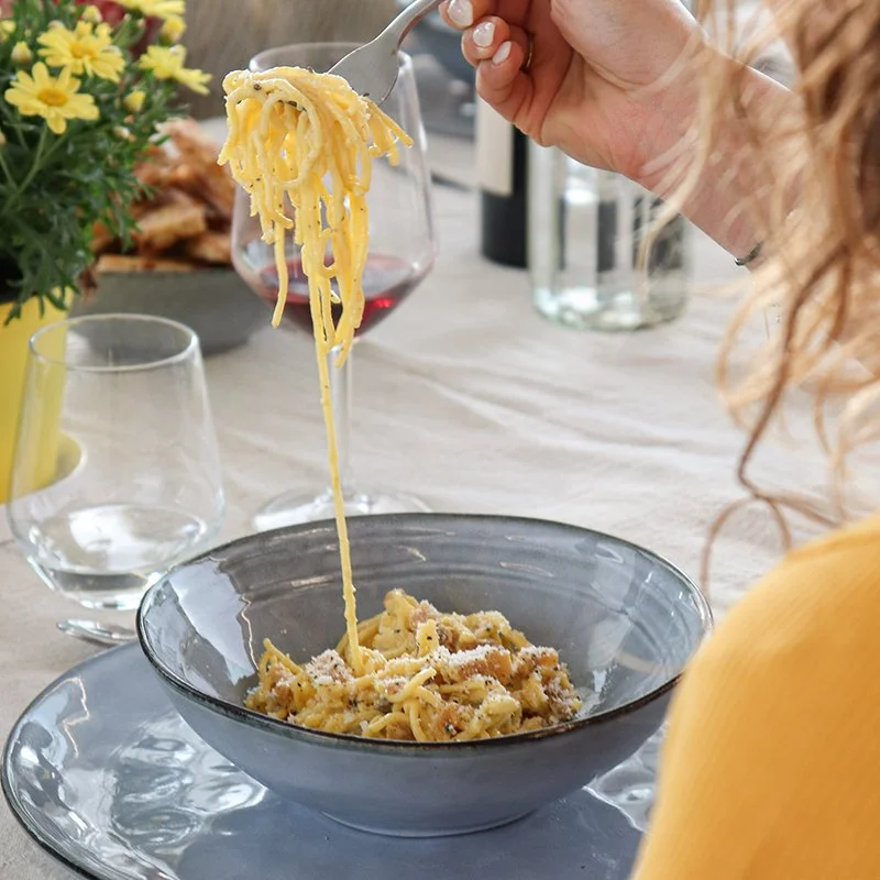 Person eating a bowl of spaghetti with grated cheese at a dining table with wine glasses, flowers, and other dishes.