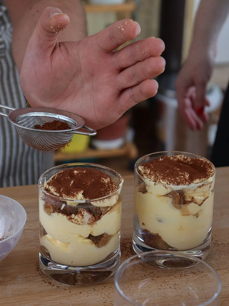 A person sprinkling cocoa powder over cups of tiramisu in glasses on a wooden surface.