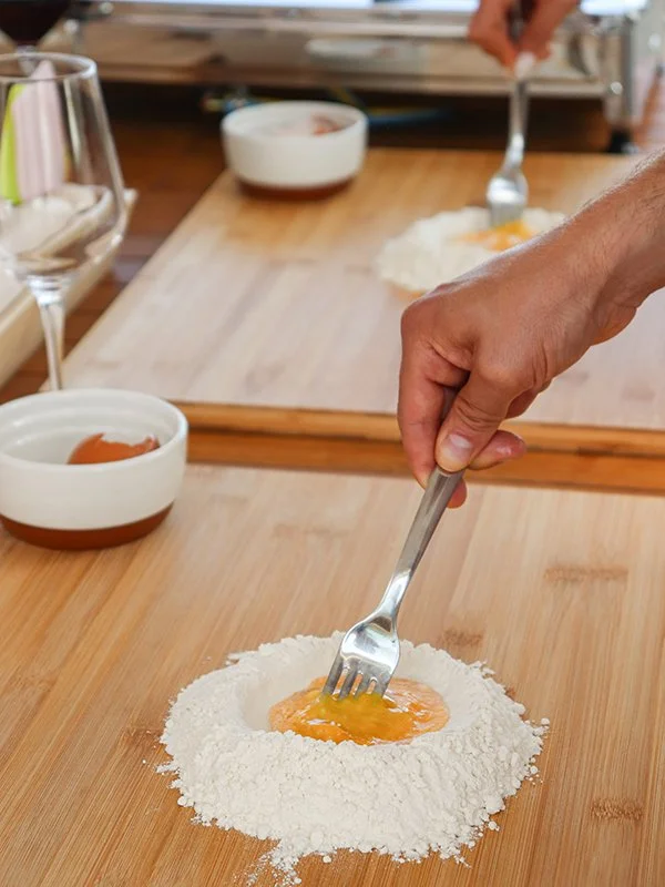Person preparing a dish by cracking an egg into flour on a wooden table, with bowls and utensils nearby.