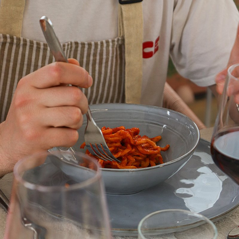 A person in a beige apron is eating spaghetti with red sauce from a gray bowl, with glasses of red wine on the table.