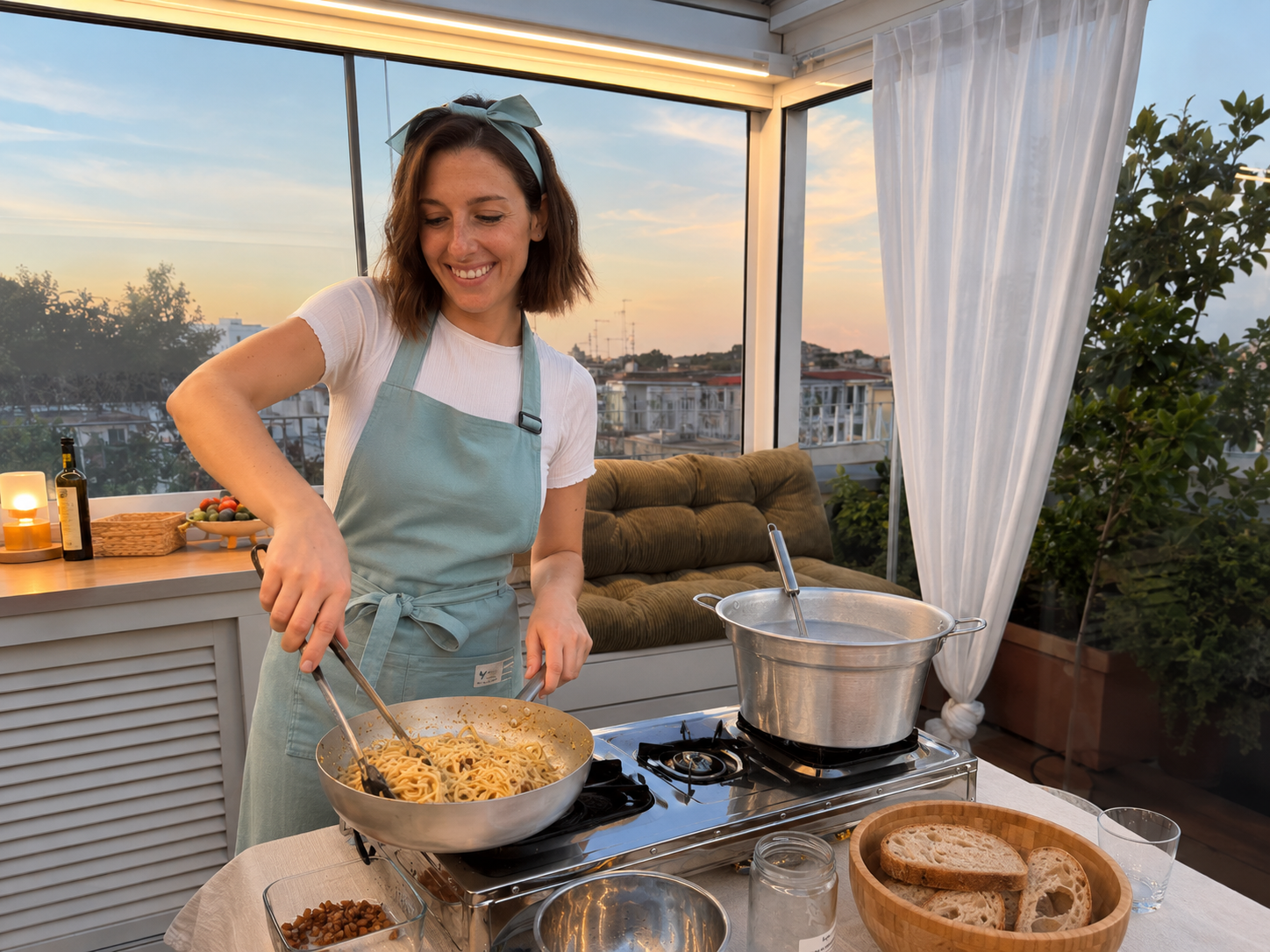 Woman cooking spaghetti pasta on a portable stove in a sunlit room with large windows and white curtains, with a view of buildings and trees outside.