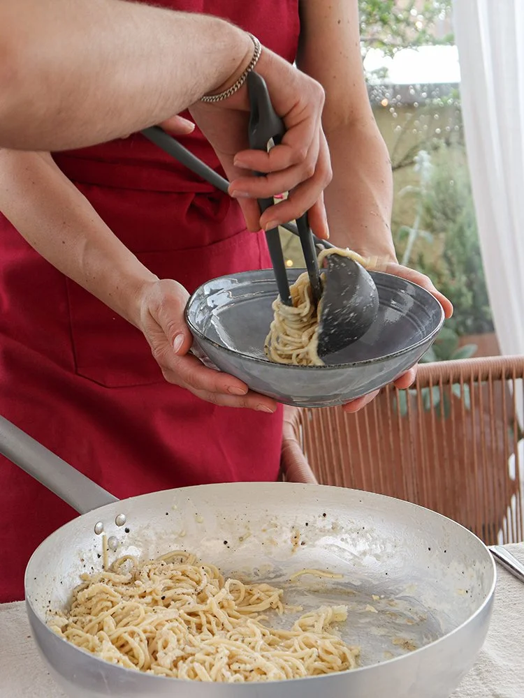 Person serving spaghetti pasta from a bowl into a plate using tongs, with more pasta in a large white mixing bowl on a table.