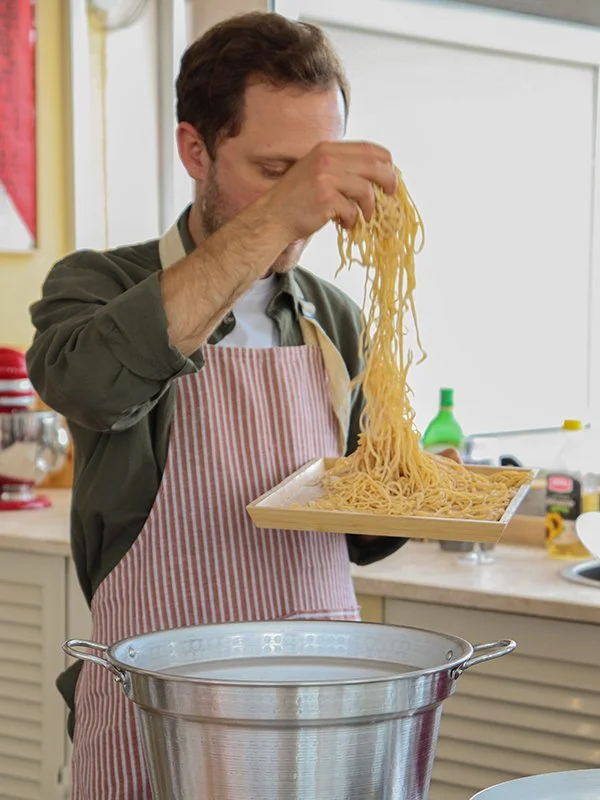A man is holding a tray of cooked spaghetti over a pot of boiling water in a kitchen.