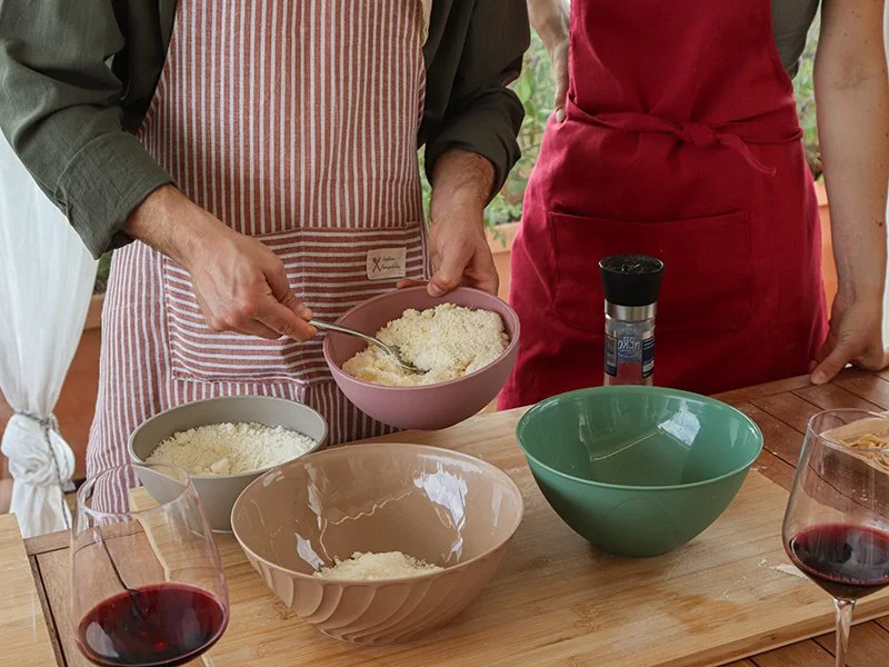 Two people standing at a wooden table, one stirring a bowl of grated cheese or rice, with multiple bowls and glasses of red wine on the table.