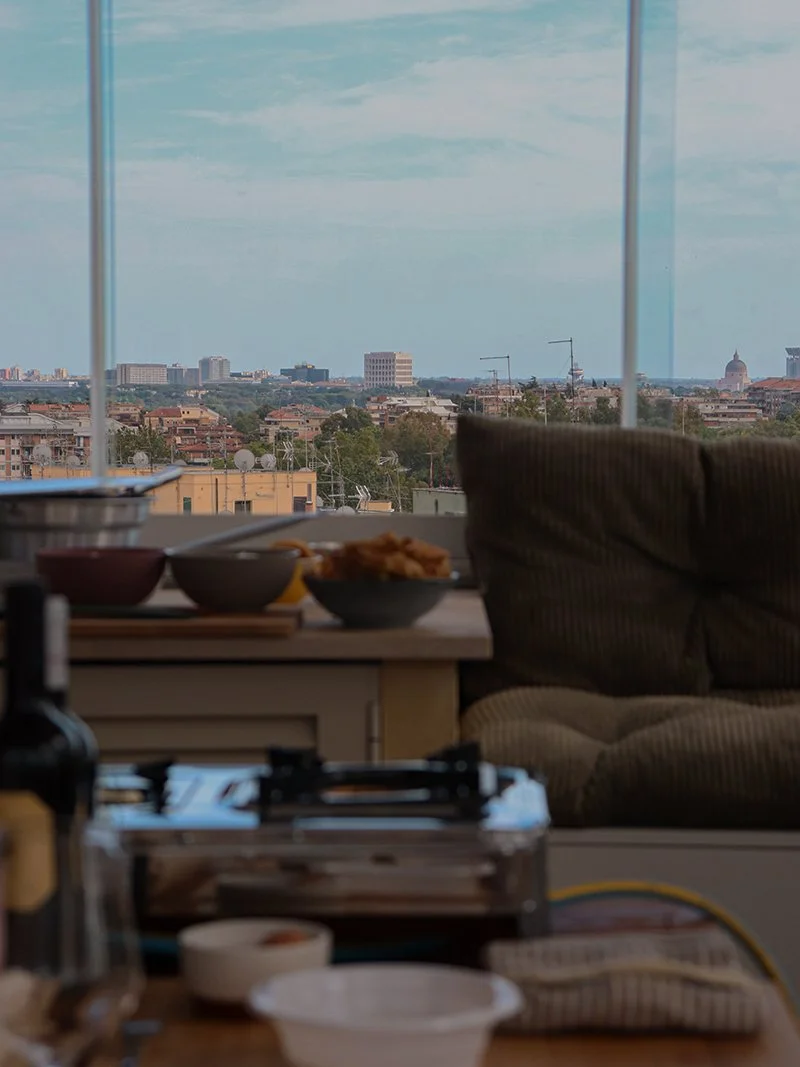A view from inside an apartment showing a cityscape through large windows, with a table in the foreground holding bowls of food and other items, and a cozy beige couch beside the window.