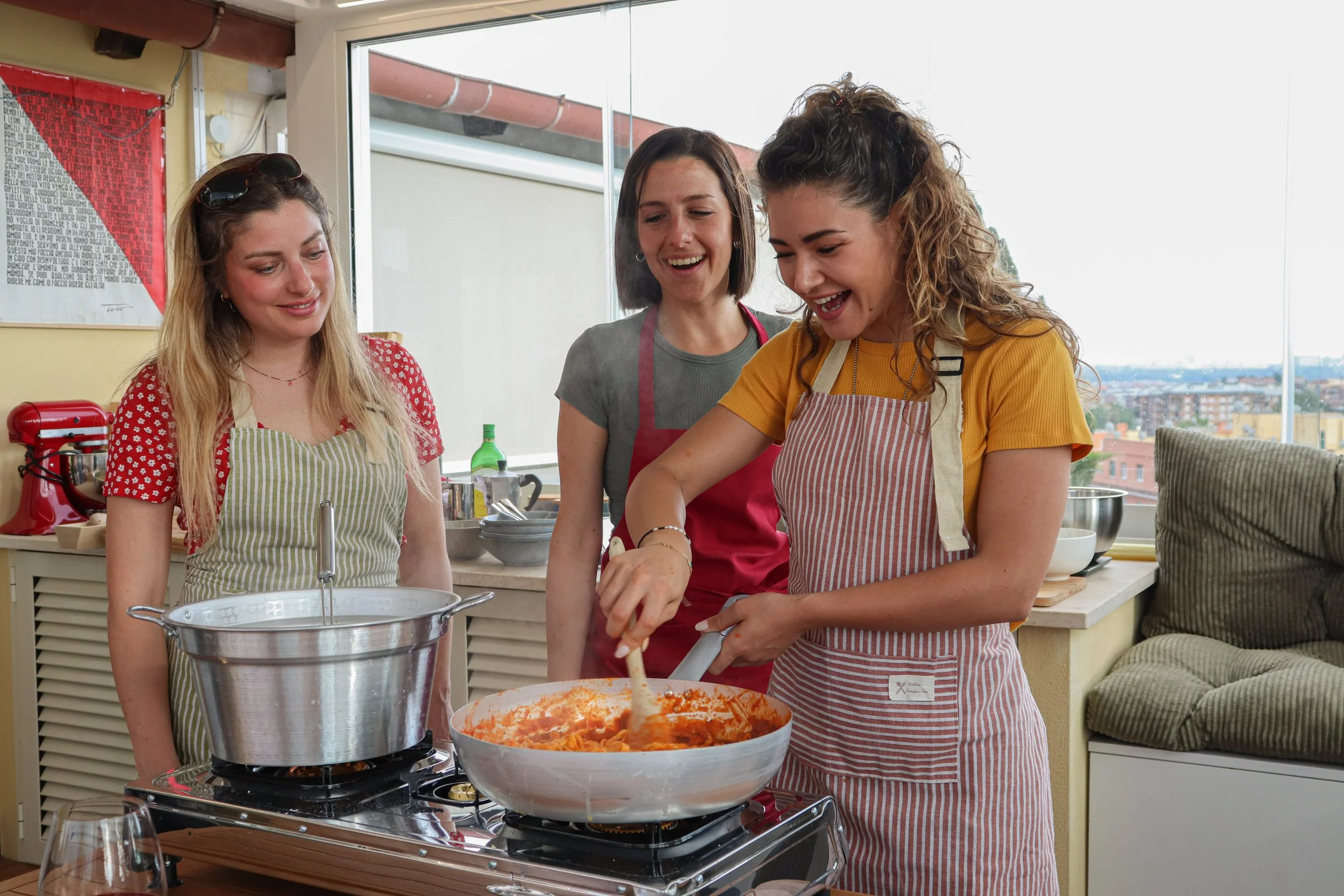 Three women cooking together in a bright kitchen, preparing a large bowl of pasta.