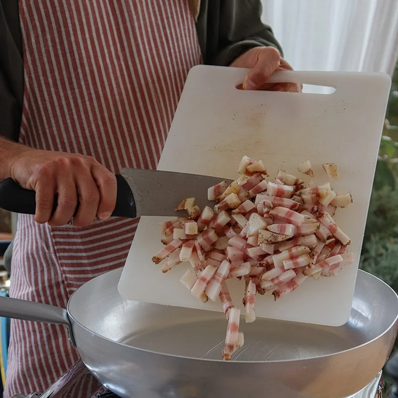 Person chopping bacon into small pieces over a large frying pan.