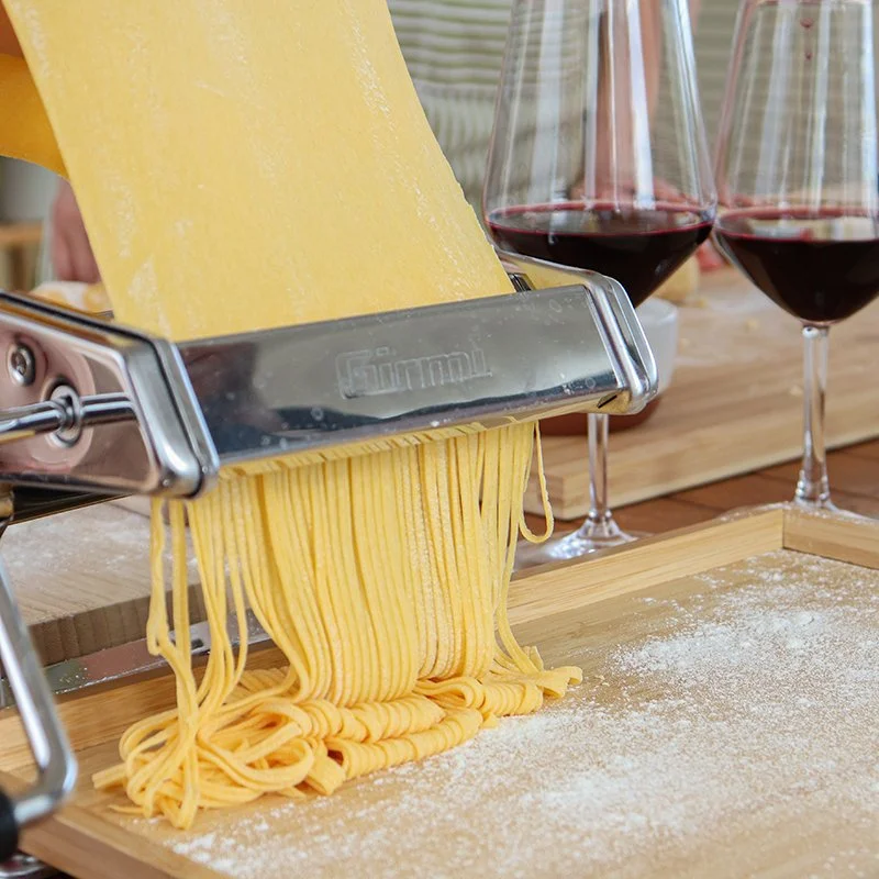 Freshly cut spaghetti being dispensed from a pasta machine with two glasses of red wine in the background on a wooden surface.