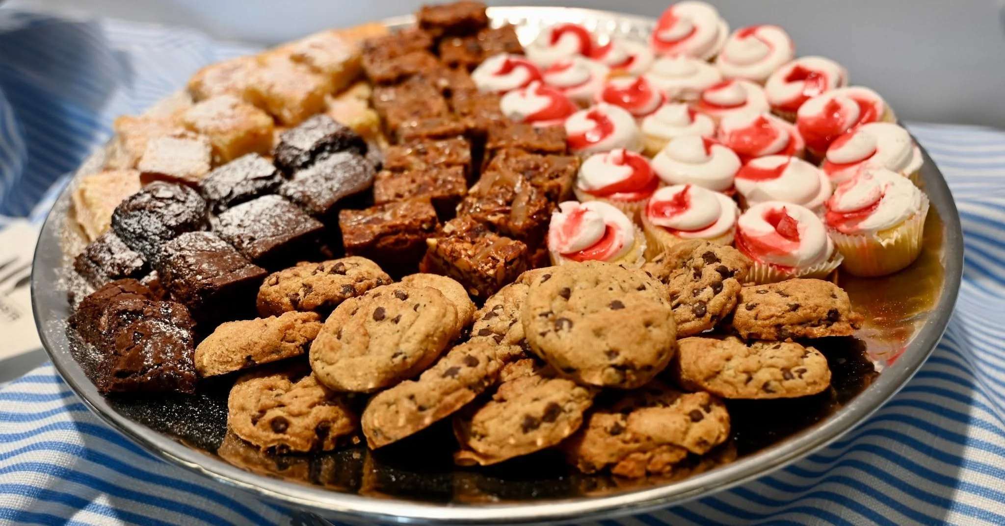 A large round platter filled with assorted cookies, brownies, and cupcakes with white and red frosting, resting on a blue and white striped cloth.