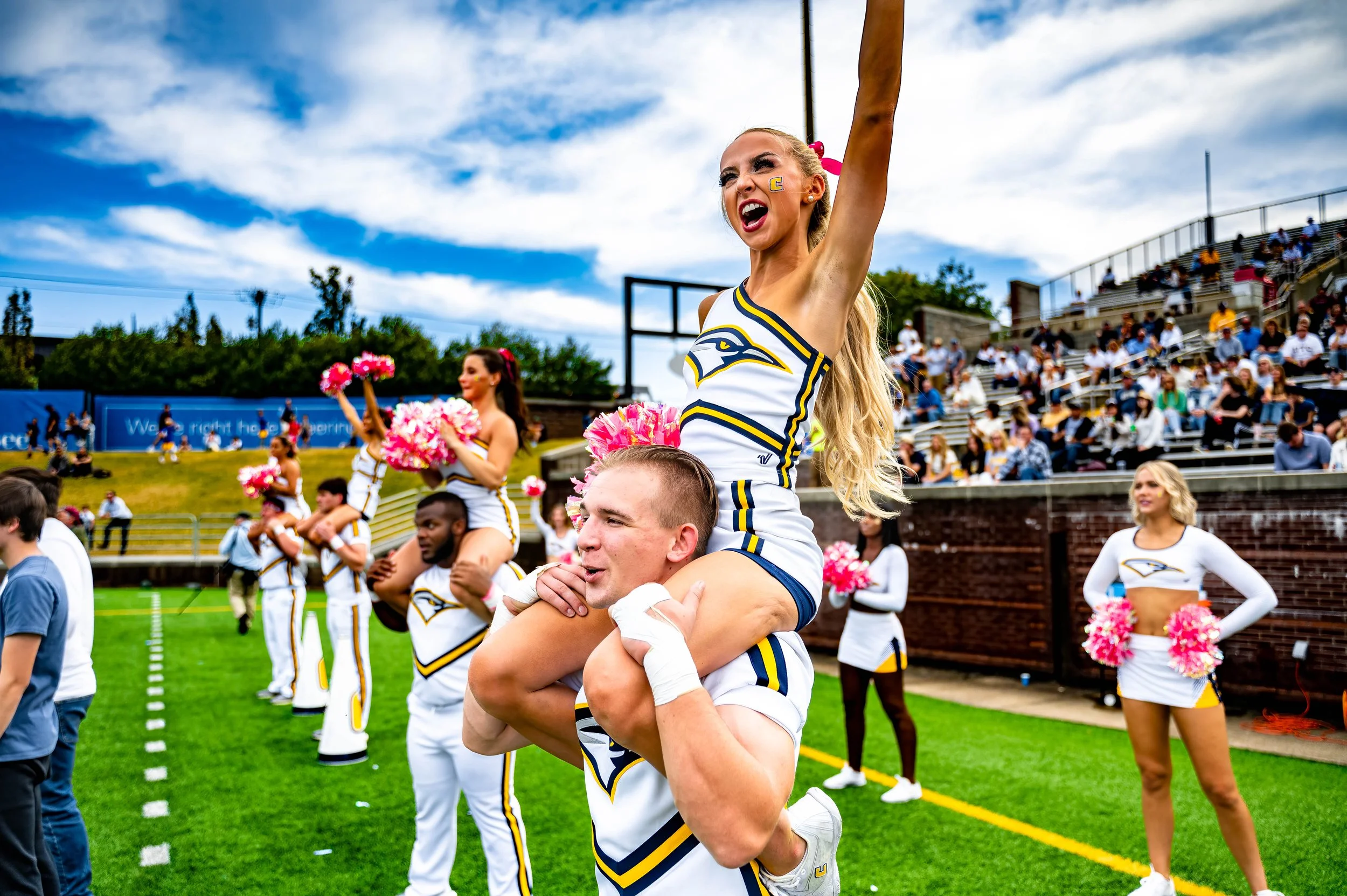 Cheerleaders performing stunts on a football field during a game, with spectators in the stands in the background.