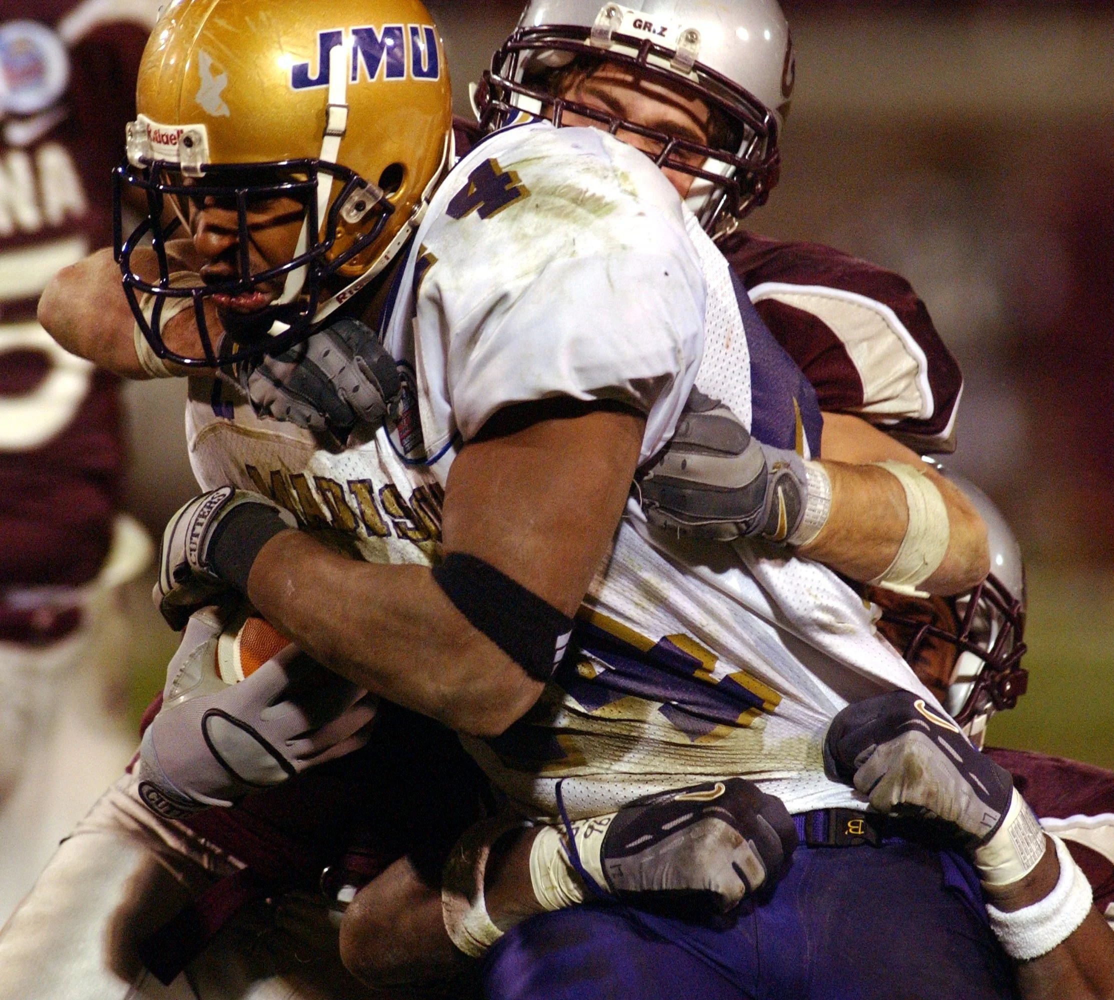 Close-up of two American football players in a tackle during a game, one wearing a gold helmet and the other wearing a silver helmet with maroon stripes, with the player in the gold helmet holding the football.