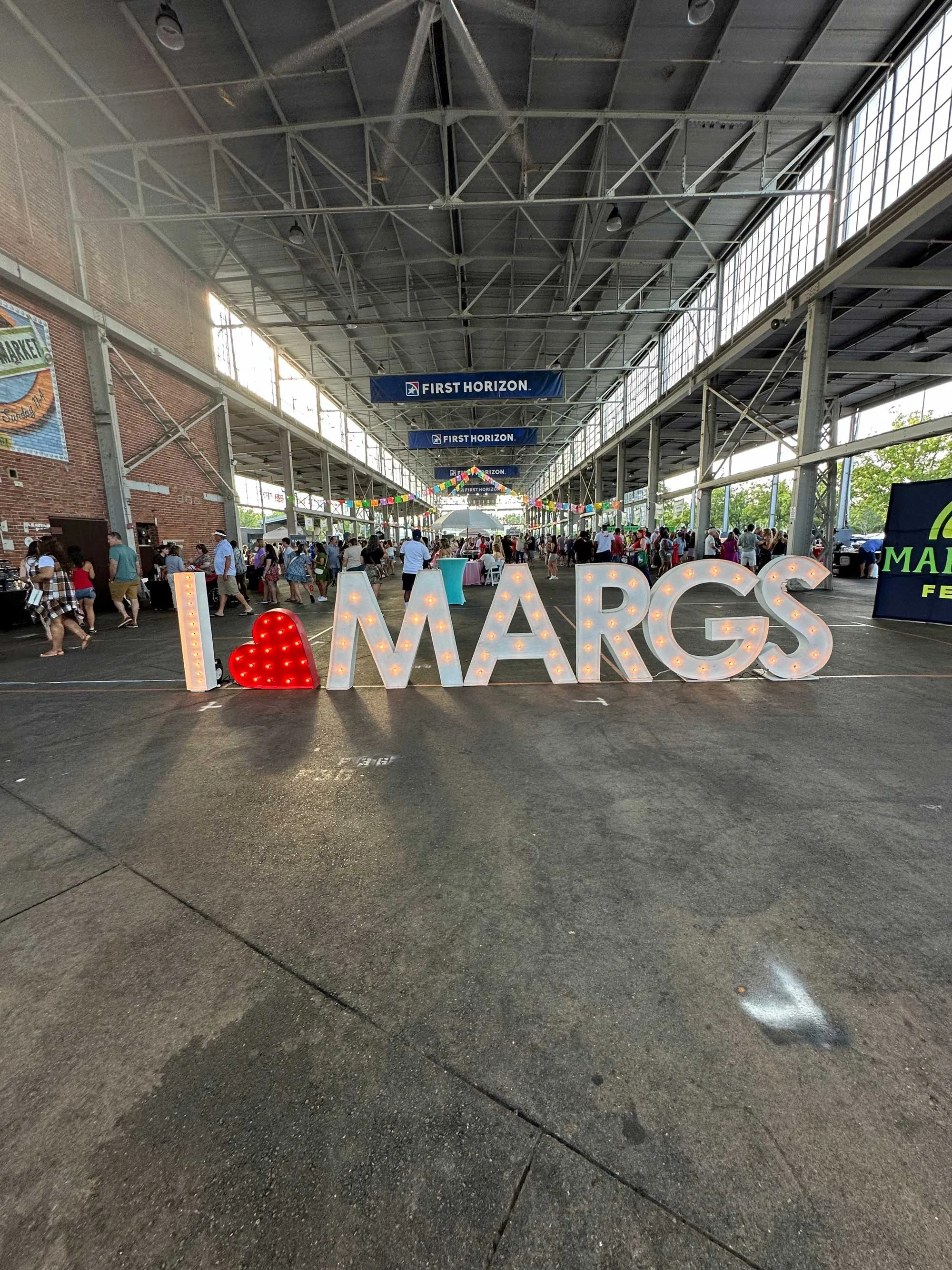 Indoor market or event space with large sign that says 'I love MARG' in front, with many people walking and shopping, decorated with colorful bunting, under a high ceiling with signs that say 'First Horizon'.