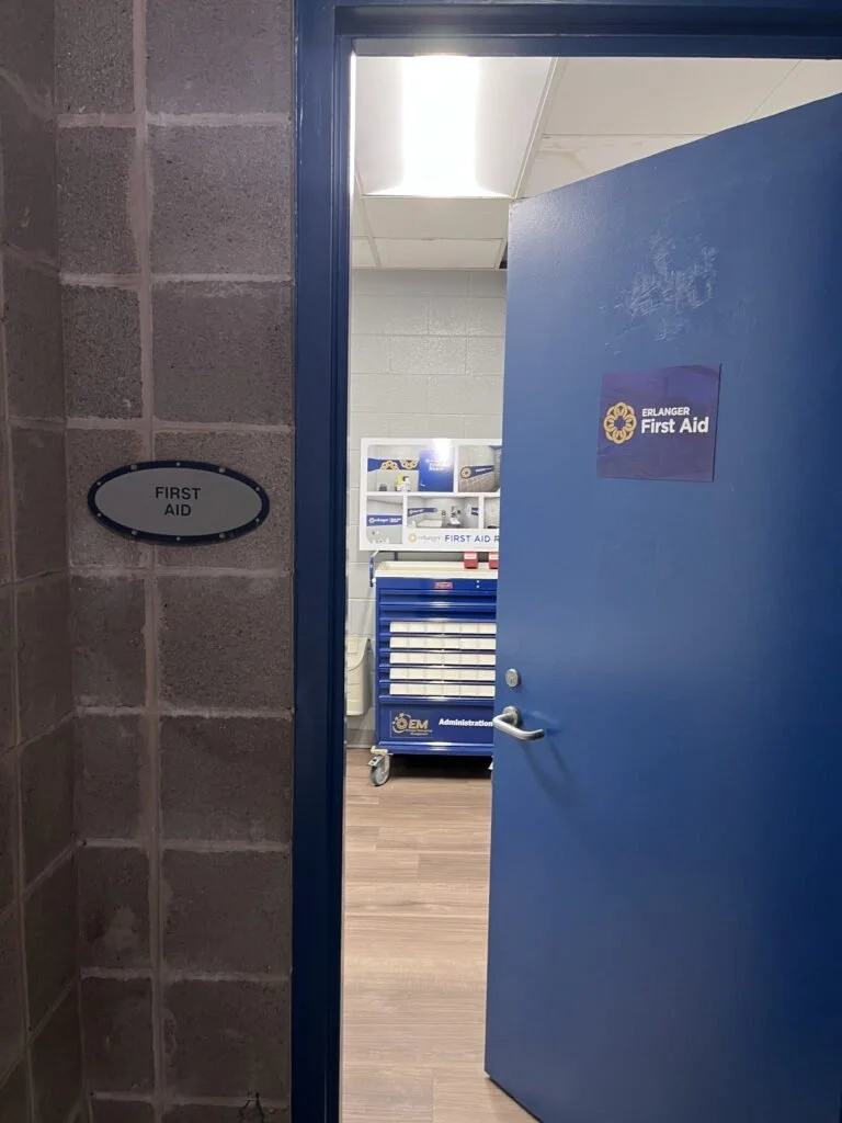 Open door leading into a first aid room, with a blue first aid sign on the door and a small oval first aid sign on the wall next to the door.