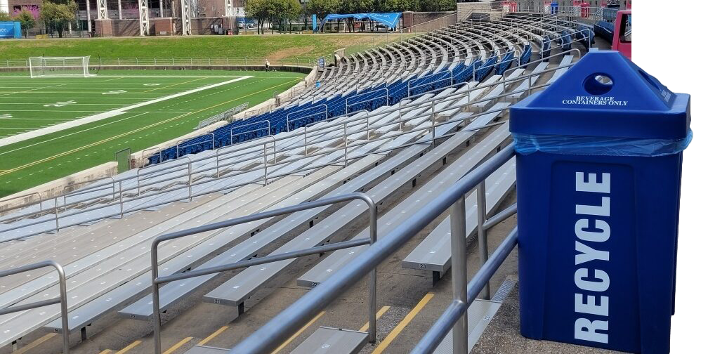 Empty football stadium bleachers with blue recycling bin labeled 'Recycle' and a sports field in the background.