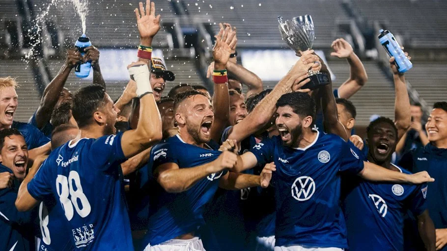 Soccer team celebrating victory, with players holding a trophy and spraying water, wearing blue jerseys with a Volkswagen logo.