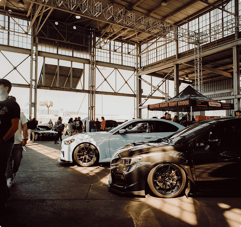 Car show inside a large industrial building with sunlight streaming in, featuring customized sports cars and people walking around.
