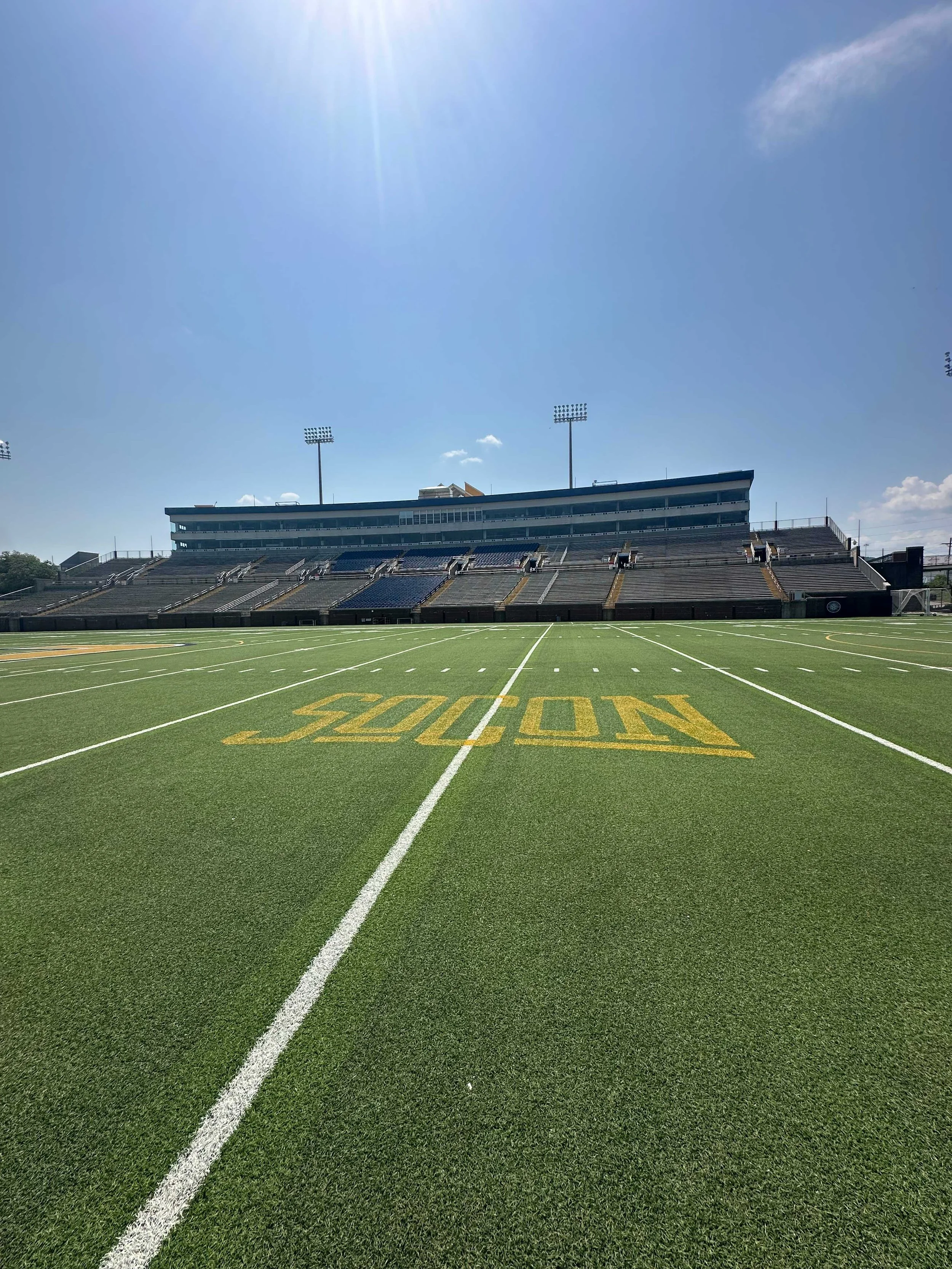 View of a football stadium field with green artificial turf, white yard lines, and a large yellow 'NO' painted on the field. The background shows stadium seating and tall light poles under a bright blue sky with some clouds and sunlight.