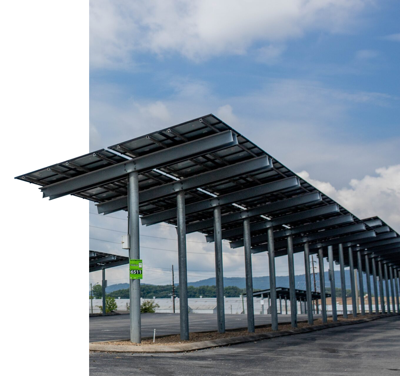Empty parking lot with solar panel-covered carport structures, green parking sign, mountains in the background, partly cloudy sky.