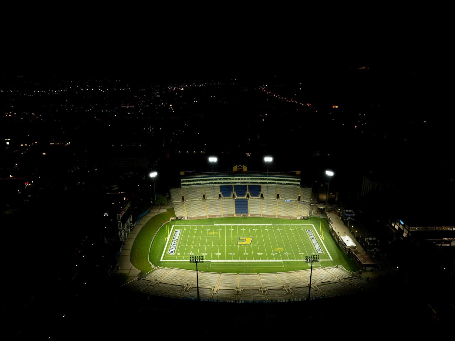 Night view of a football stadium illuminated by floodlights, surrounded by an empty seating area, with city lights in the distance.