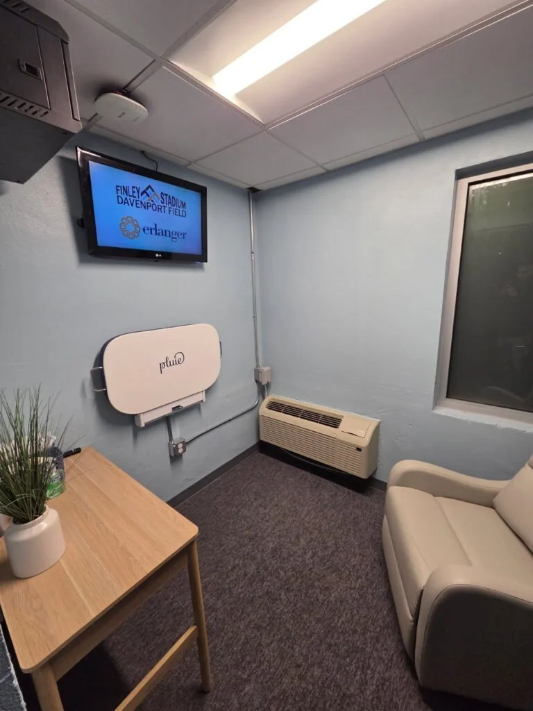 Small room with a beige chair, a wooden table with a plant, a wall-mounted TV displaying 'Finley Stadium Davenport Field Erlanger,' a wall heater, and a window with a view outside.