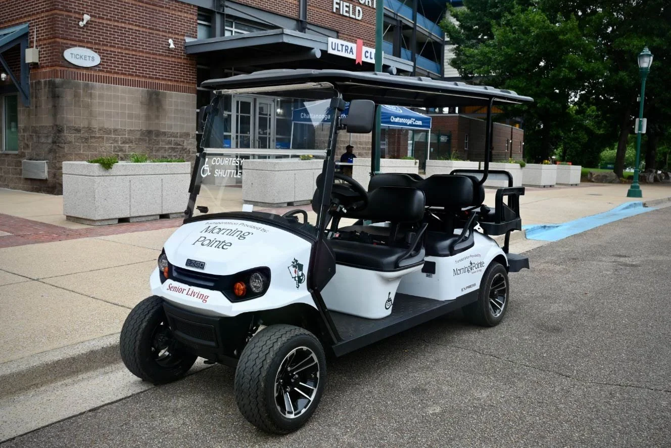 A white golf cart with the words 'Morning Pointe' and 'Senior Living' is parked on a sidewalk outside a brick building with large planters. The cart has four seats and a roof, and there are signs indicating volunteer shuttle service.