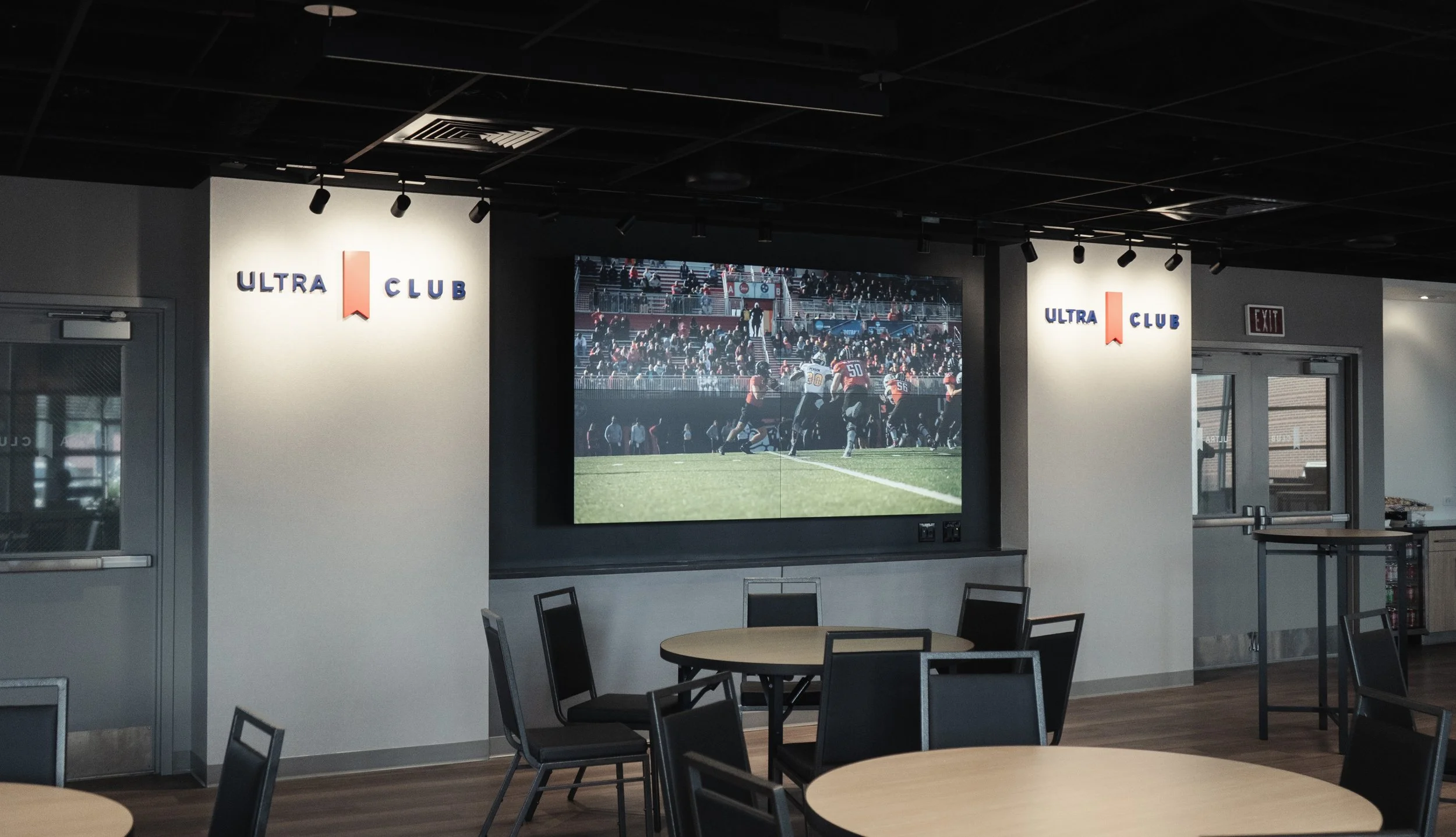 Indoor area with round tables and chairs, large screen showing a football game, illuminated signs for Ultra Club, and glass doors at the back.