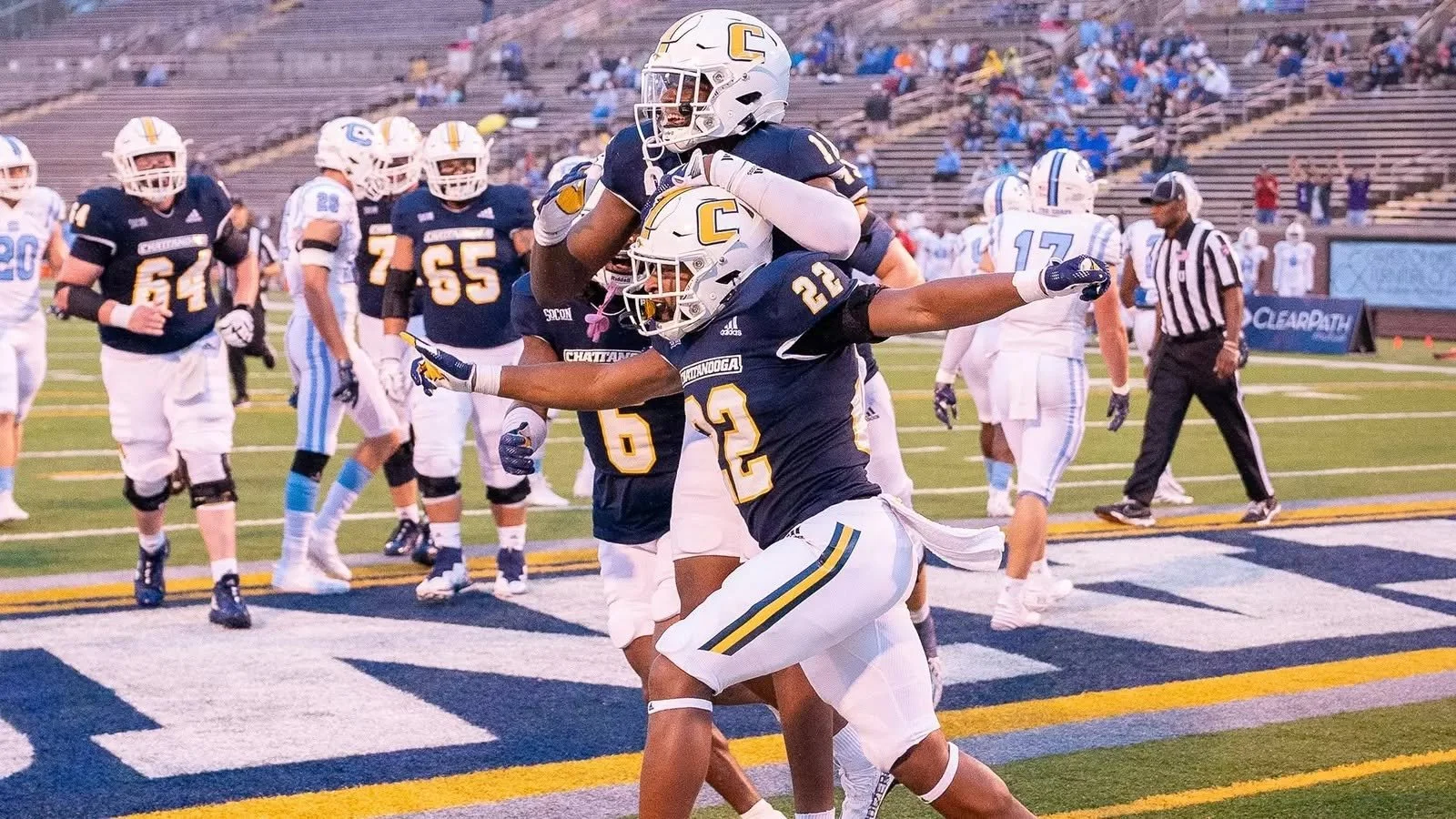 Two football players in blue uniforms celebrate a touchdown on the field, with one player jumping onto the other's back, surrounded by teammates and an official.