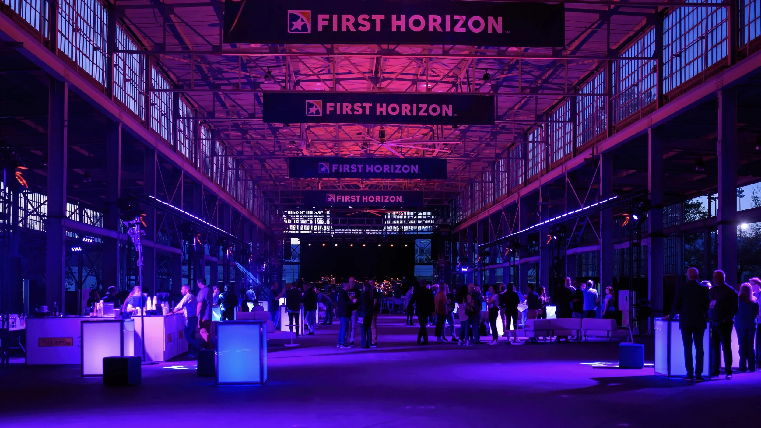 Indoor event space with purple and pink lighting, banners displaying 'First Horizon' hanging from the ceiling, and people standing and sitting at tables.