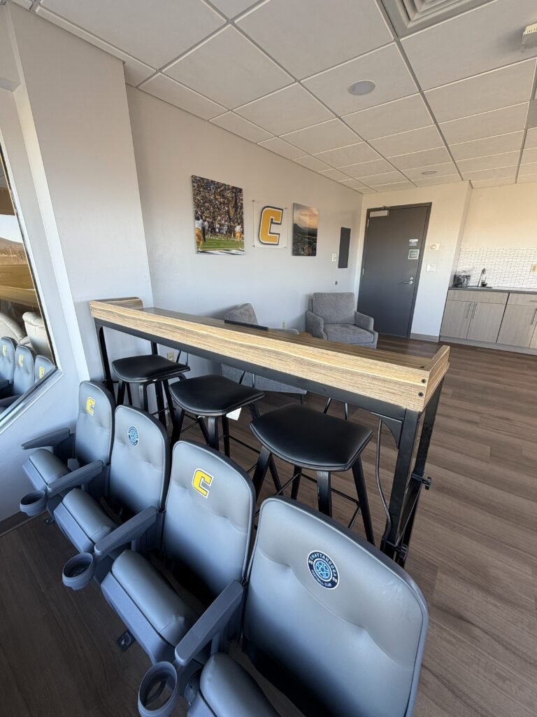 Indoor lounge area with a row of grey stadium-style seats with a yellow letter 'C' on the headrests, a high wooden table with black stools, grey armchairs, and artwork on the wall including a football scene, a 'C' letter, and a landscape. There is a kitchen counter in the background.