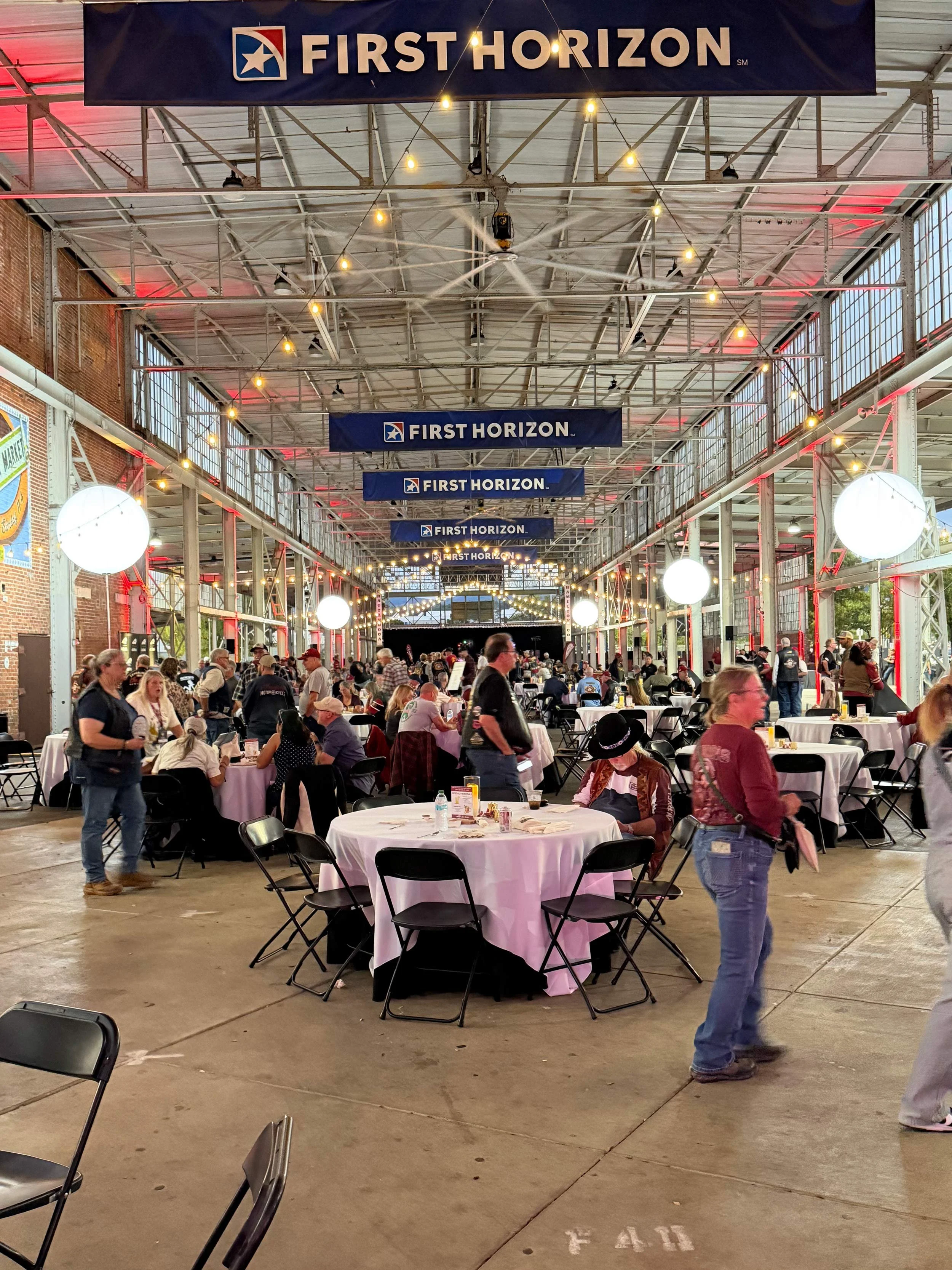 A large indoor event with round tables covered in pink tablecloths and surrounded by black chairs, filled with people dining and socializing. The space has high ceilings with metal beams and windows, decorated with banners that read 'First Horizon.' String lights and large spherical lamps hang from the ceiling, creating a festive atmosphere.