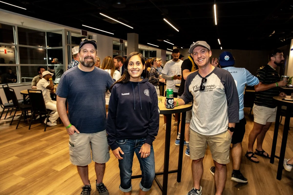 Three people stand together in a social gathering, smiling at the camera. The setting appears to be indoors with other people socializing in the background. The person in the middle is wearing a dark hoodie, while the two men on either side are in casual t-shirts and shorts, with one holding a drink.