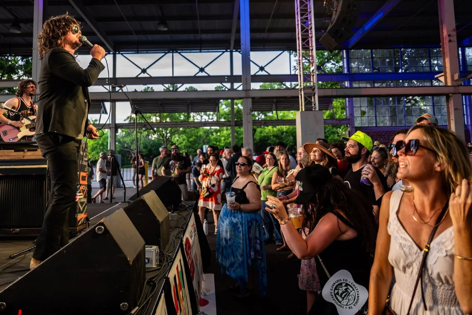 Musicians perform on stage at an outdoor concert, with an audience enjoying the show under a partially covered structure surrounded by greenery.
