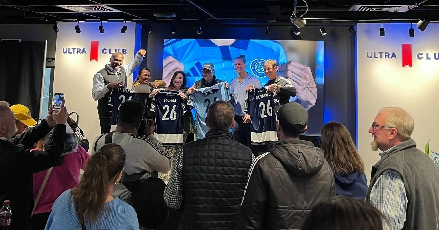 People on stage holding soccer jerseys during an event, with an audience watching and taking photos, and a large screen with an athlete's hands signing a jersey in the background, in a room decorated with 'ULTRA CLUB' banners.