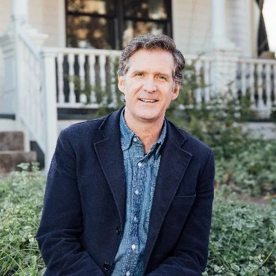 A man in a blue blazer and blue shirt standing outdoors in front of a house with a white porch railing.