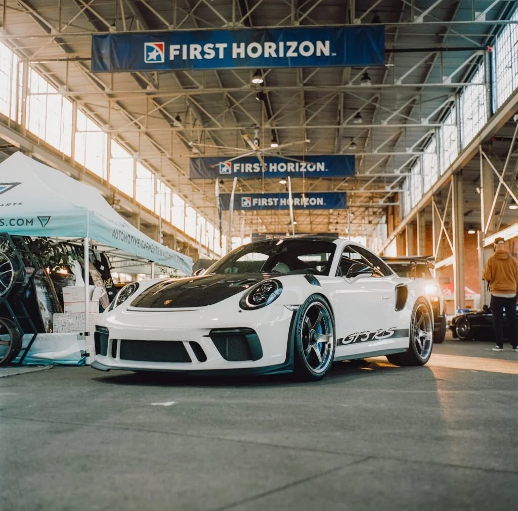 A white Porsche 911 GT3 RS with black accents and racing wheels on display at an indoor car show, with banners reading 'FIRST HORIZON' hanging above the vehicle.
