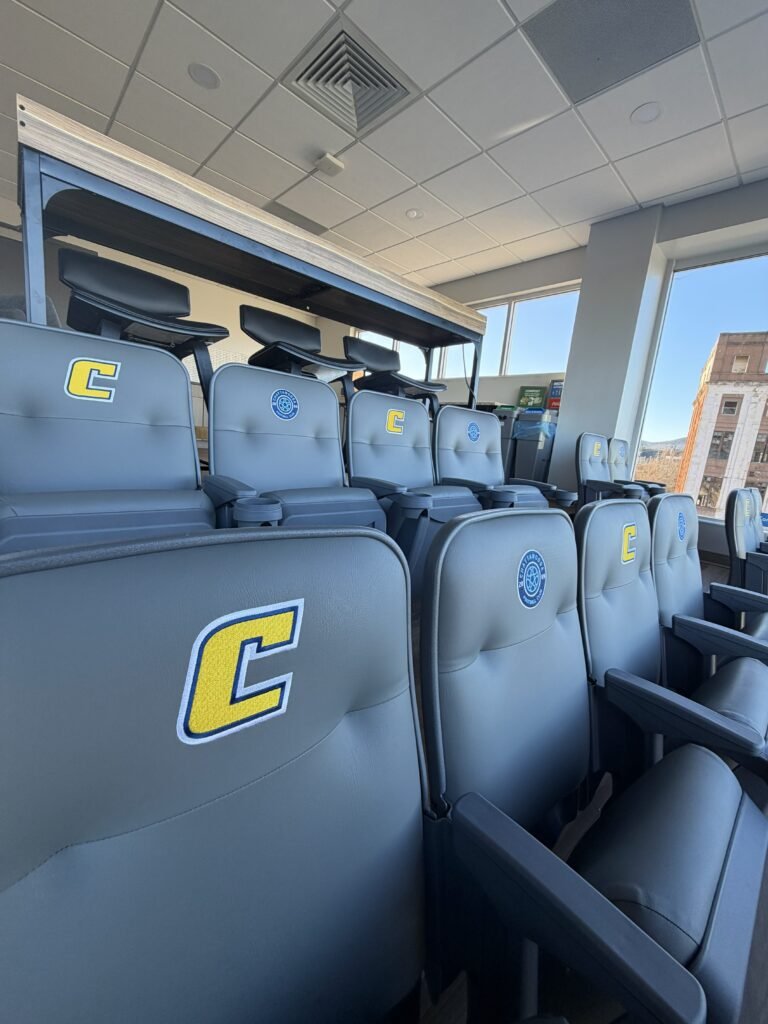 View of empty stadium seats with a letter C patch and a circular emblem on the headrests, near large windows showing a cityscape outside.