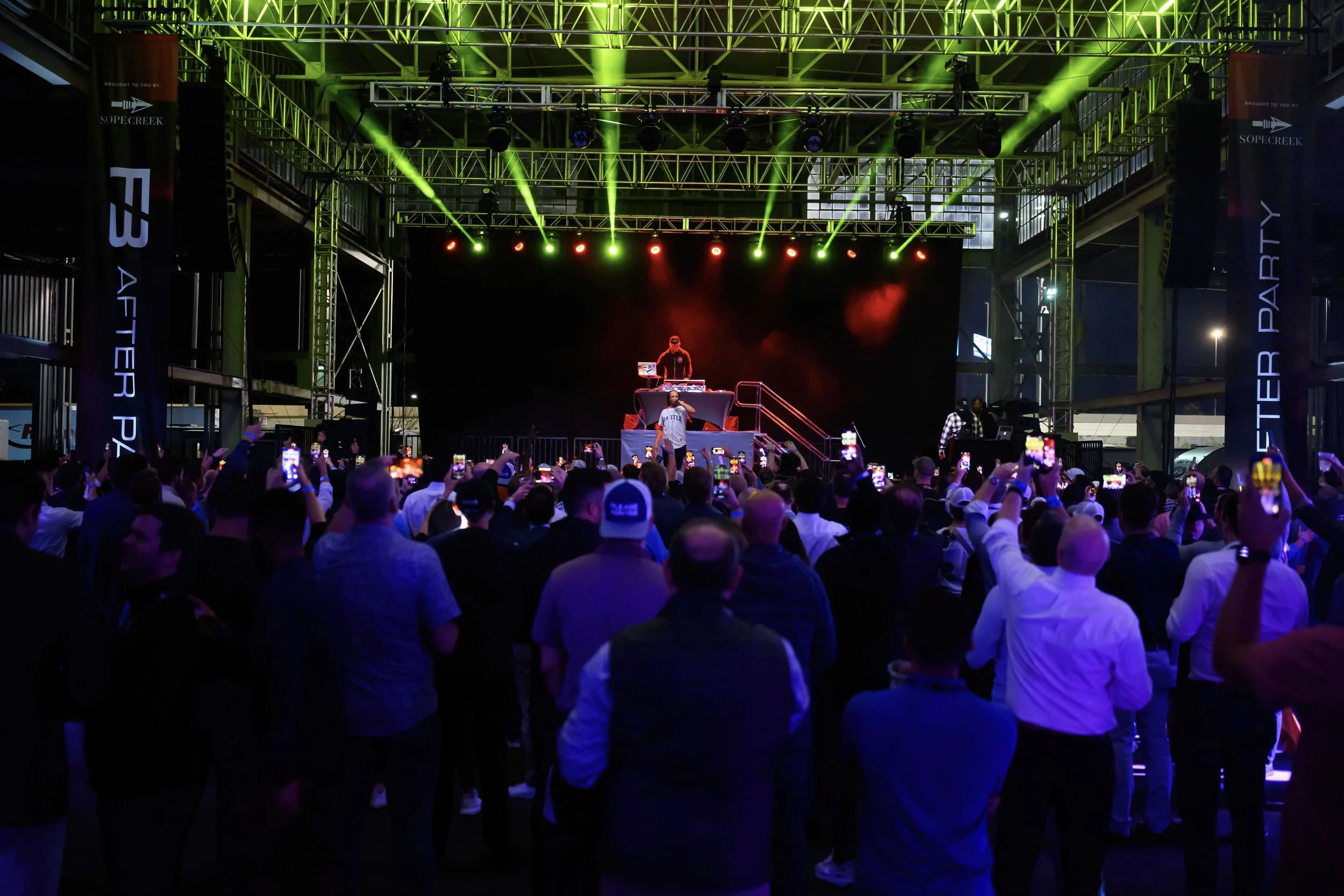 A large crowd attending an outdoor concert or event with a stage featuring green and red lighting, a DJ performing at a booth, and many audience members taking photos with their phones.