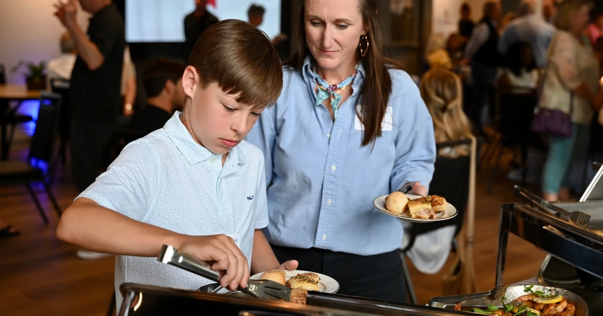 A woman and a young boy at a buffet table, with the woman holding a plate of food and the boy serving himself, in a crowded indoor setting.