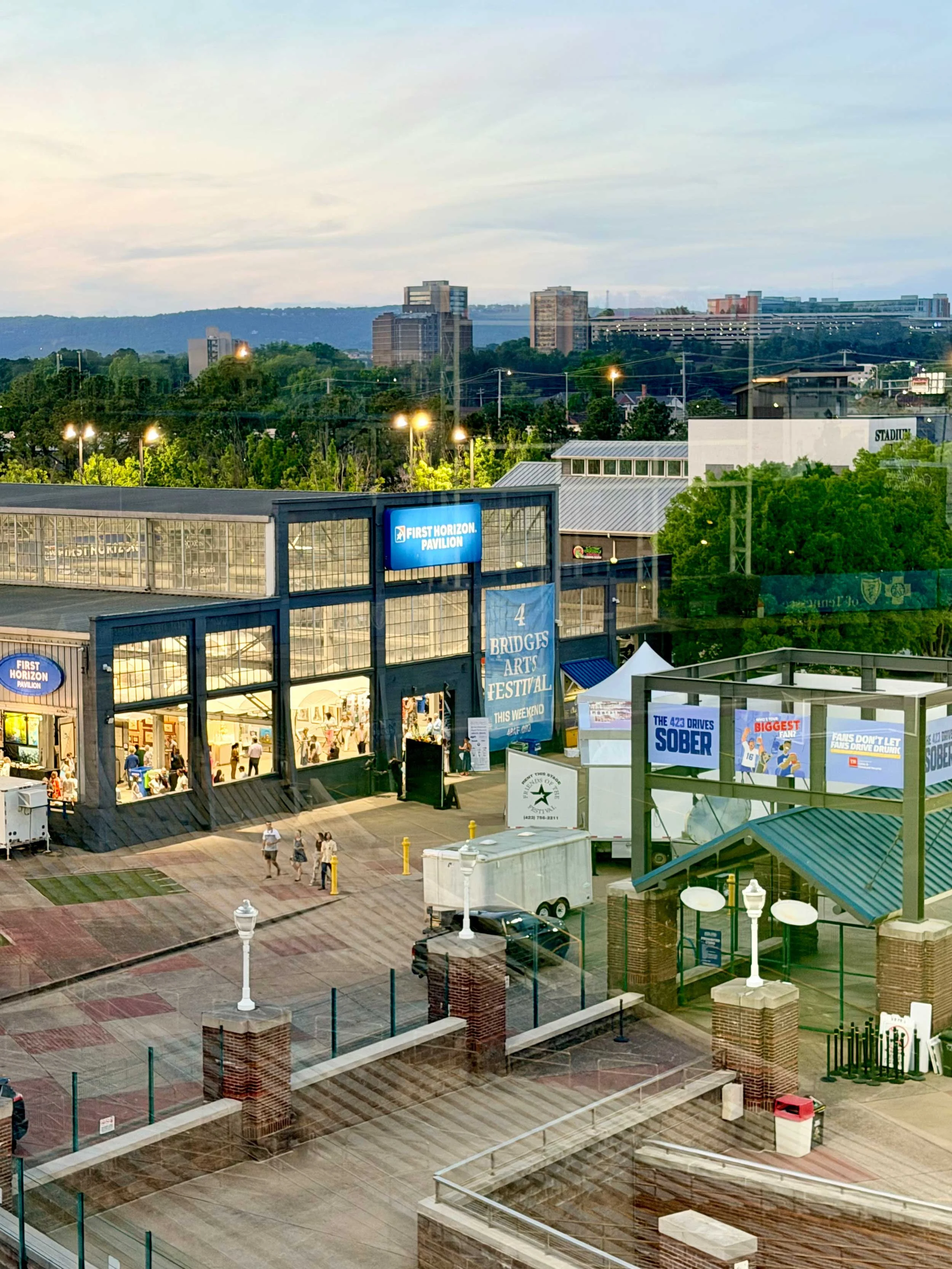 A view of outdoor event space with people walking, tents, banners, and buildings in the distance at dusk.