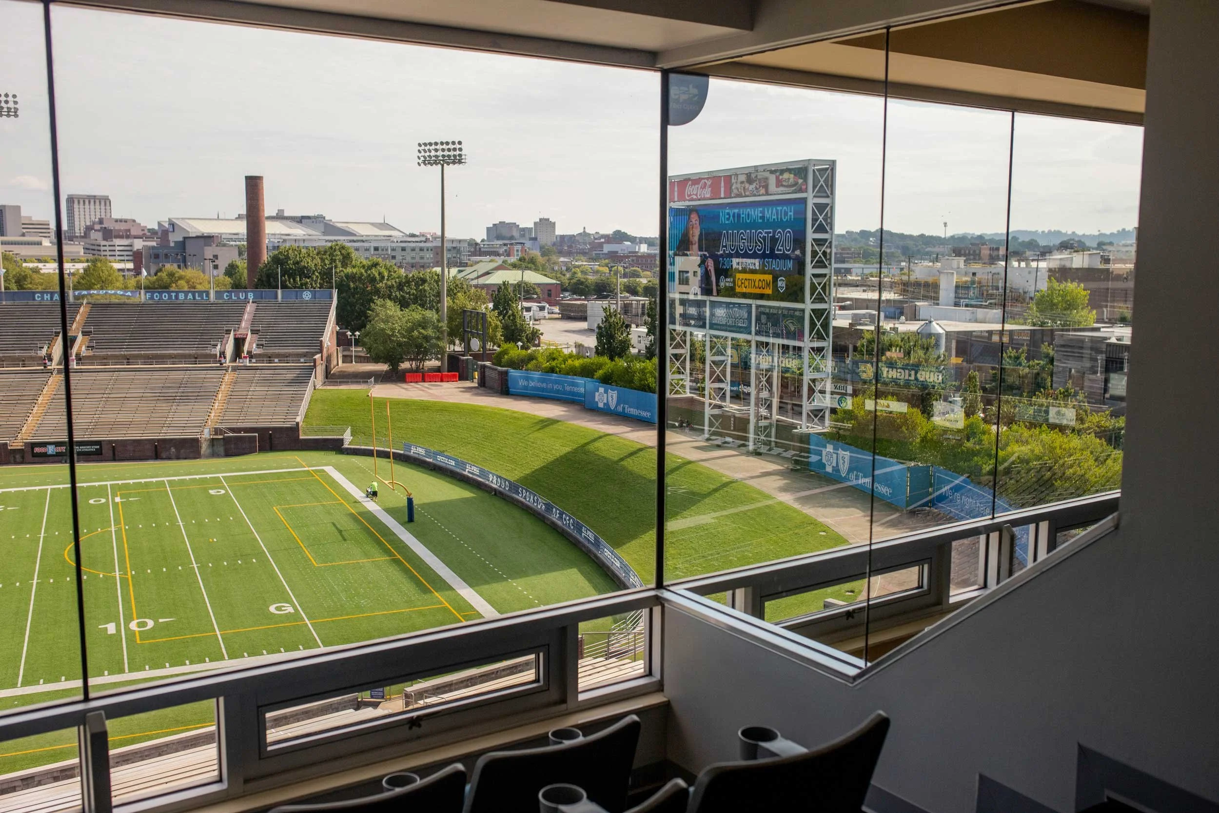 View of a football stadium seen through large windows, with empty stadium seats, a scoreboard advertising an upcoming game on August 20, and surrounding cityscape in the background.