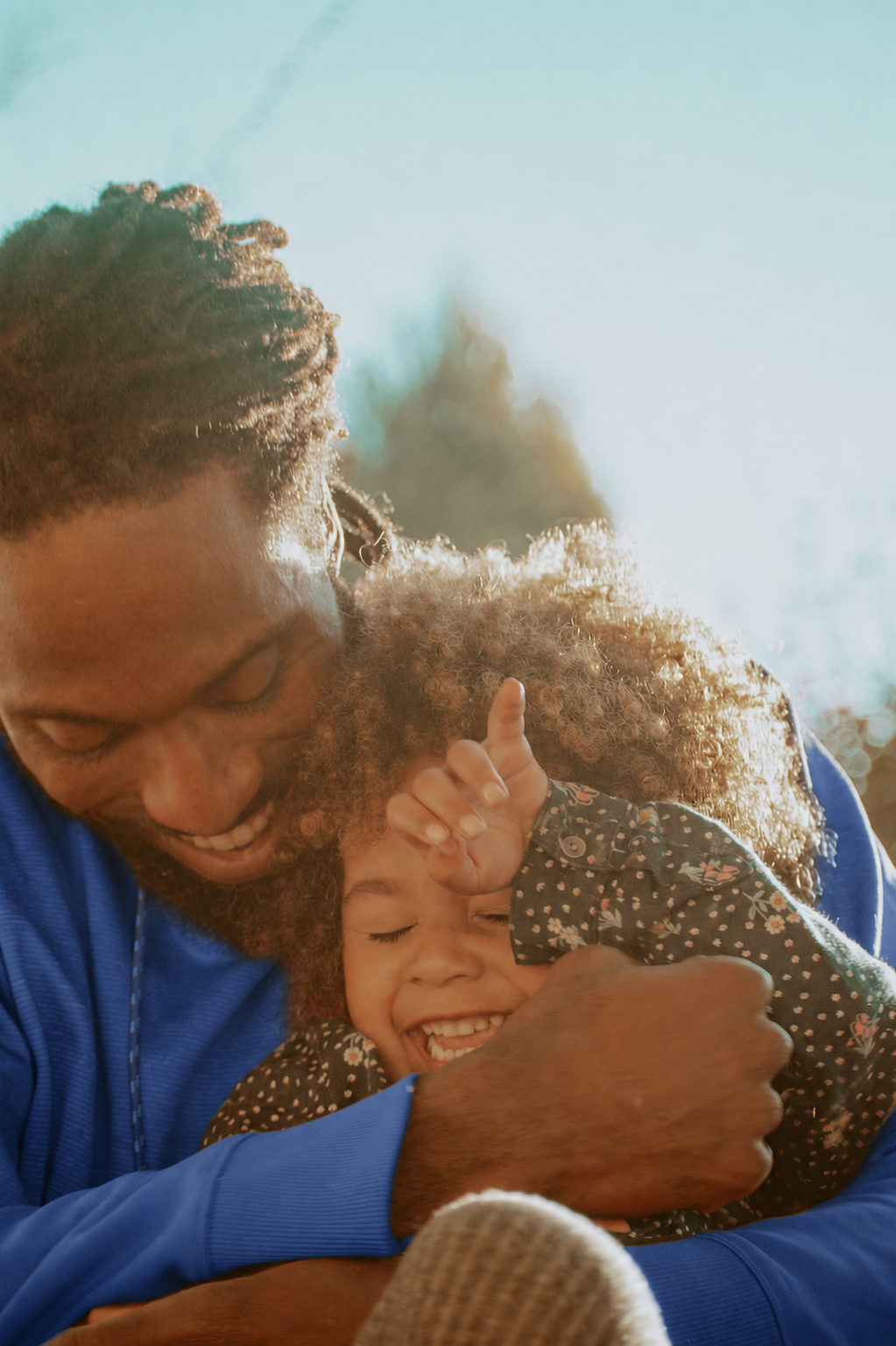 A man and a young girl sharing a joyful embrace outdoors, smiling with their eyes closed, the girl playfully touching her forehead with her hand, sunlight illuminating their faces and curly hair.