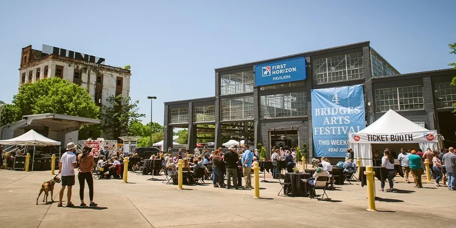 Crowd gathered outside a building for the Bridges Arts Festival, with tents and activity booths, on a sunny day.