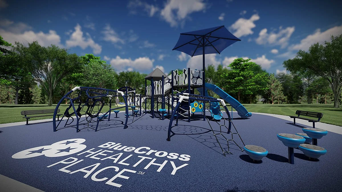 A playground with climbing structures, slides, benches, and an umbrella, on a rubberized surface with Blue Cross Blue Shield of Arizona Healthy Place written on it, surrounded by trees and a blue sky with clouds.