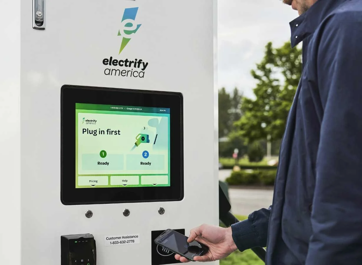 A person using a smartphone interacts with an electric vehicle charging station labeled 'electrify america' outdoors. The station has a screen displaying instructions to 'Plug in first' with two steps marked 'Ready.' The background shows trees and a partly cloudy sky.