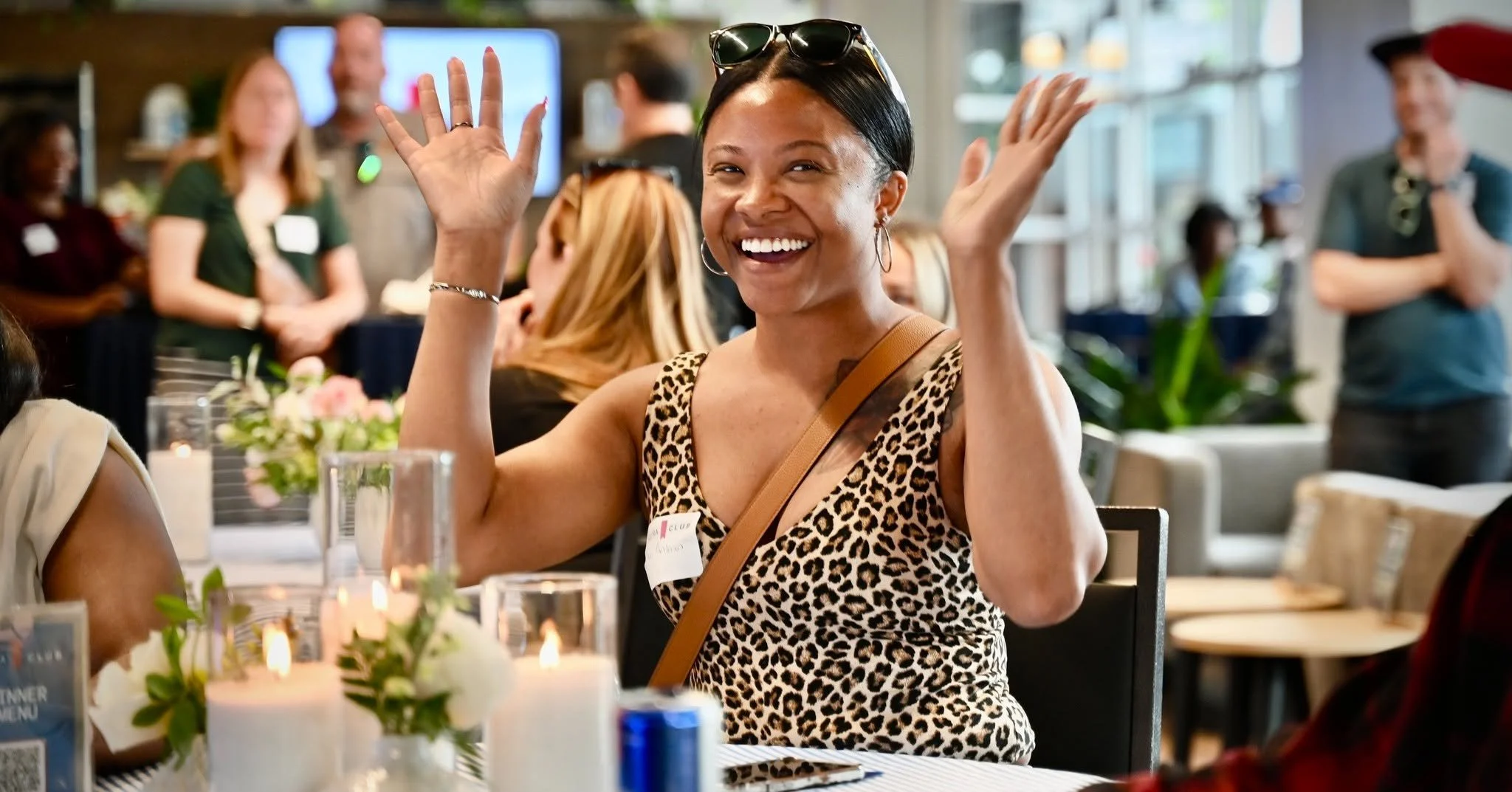 Woman with sunglasses and earrings smiling and raising her hands, sitting at a table in a lively indoor gathering, with other people in the background.