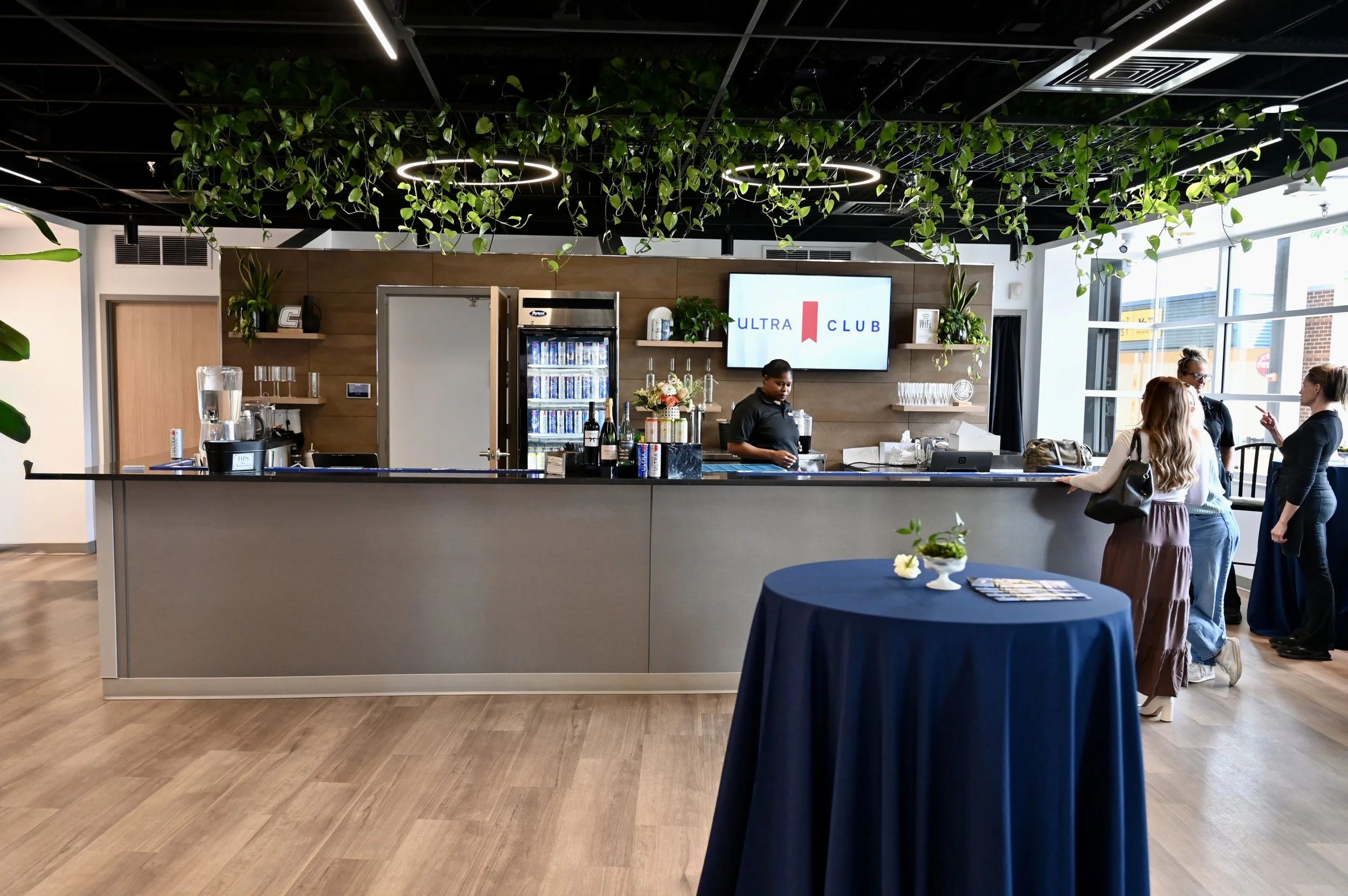 Modern indoor lounge or event space with a bar counter, greenery hanging from the ceiling, and a large screen displaying 'ULTRA CLUB'. Four women are engaged in conversation near the bar, and there is a round table with a blue tablecloth in the foreground.