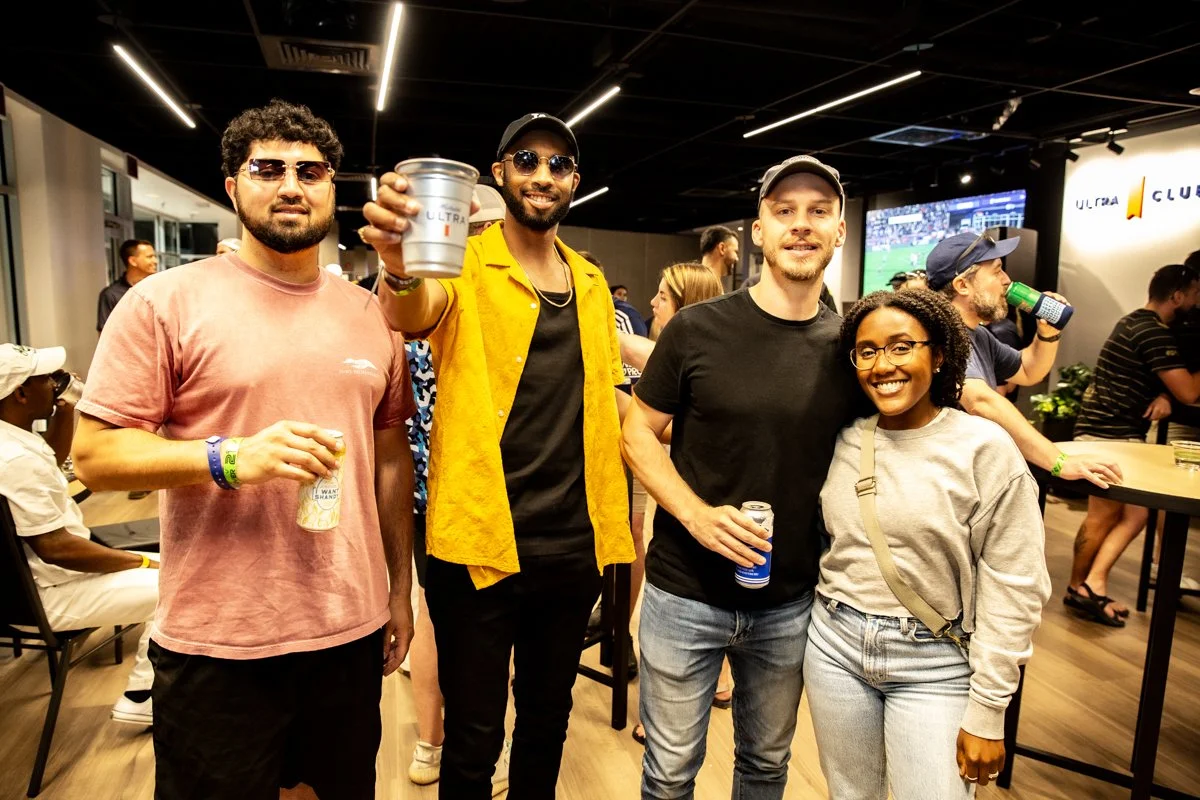 Four people smiling at a social gathering, holding drinks, with others in the background watching a sporting event on TV.
